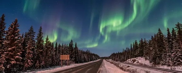Une route du Nord et une voie ferrée traversant une forêt d'épinettes sous une aurore boréale verte