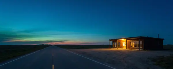 Une route des Prairies à deux voies à l'heure bleue, avec un bâtiment bas en bord de route éclairé de l'intérieur